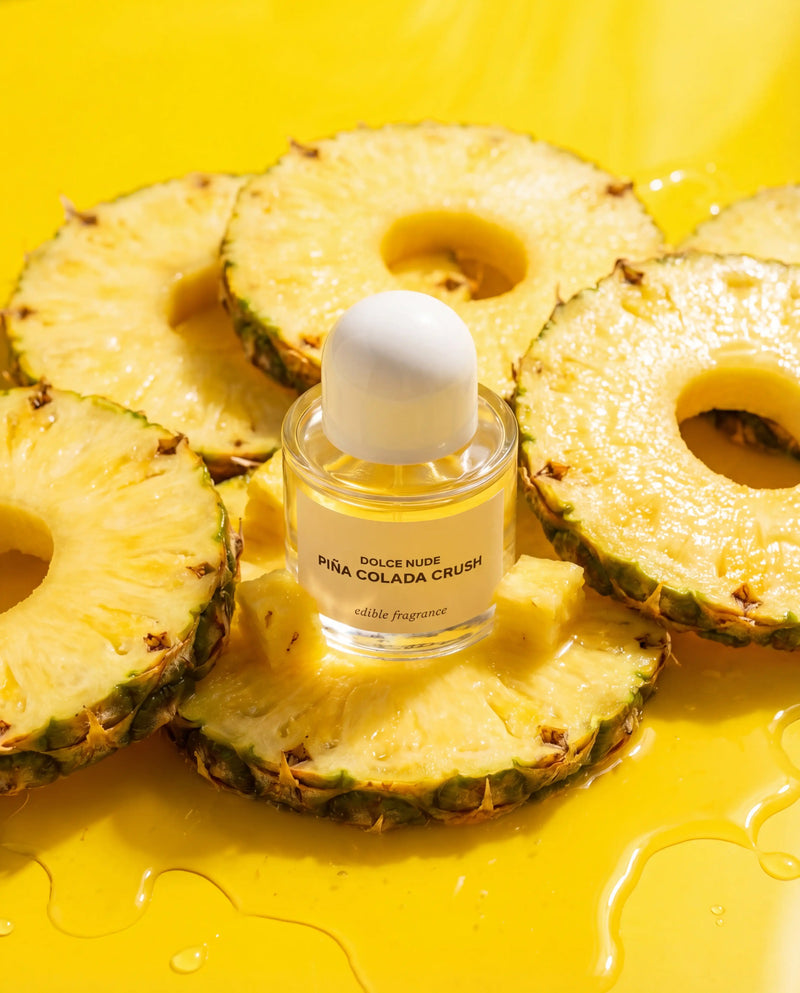 DOLCE NUDE Piña Colada Crush edible perfume bottle floating amid fresh pineapple slices on bright yellow